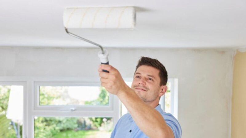 Man Painting Ceiling In Room Of House With Paint Roller